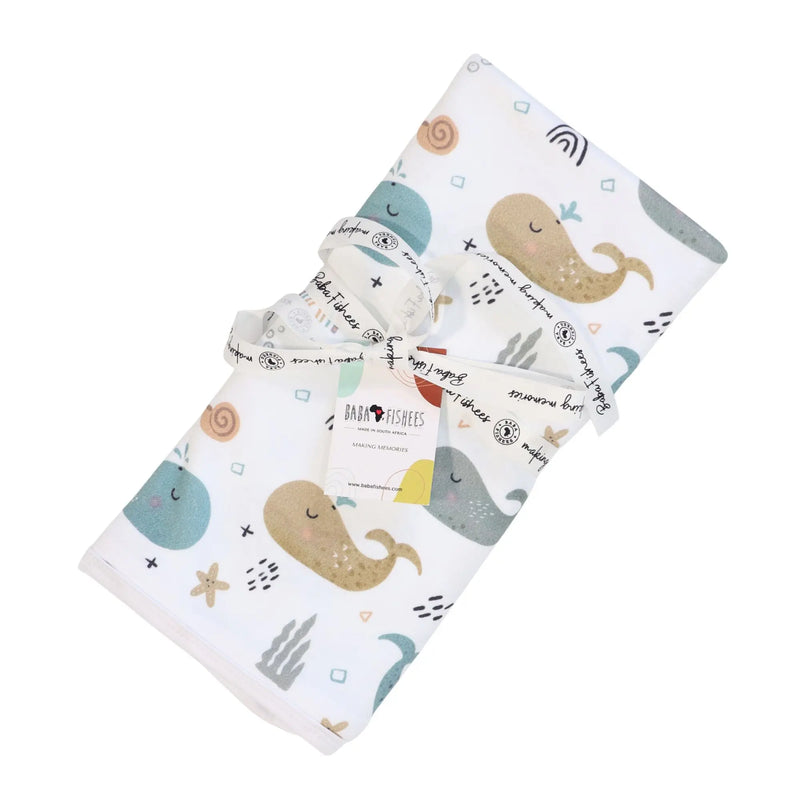 Baby blanket with whales on white background, featuring teal, sandy brown, and grey whale print rolled up with a ribbon.