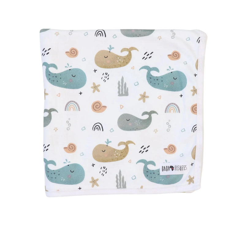 Baby blanket with whales on white background, featuring teal, sandy brown, and grey whale print folded up showing the Bab Fishees tag.