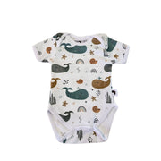 A front view of a short-sleeve white baby bodysuit with an all-over whimsical print of whales, starfish, rainbows, and seashells in mustard, blues and greys.