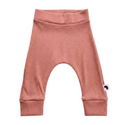 A pair of pink ribbed knit baby pants with an arched leg design, ribbed waistband, and ribbed ankle cuffs, featuring a small white tag with a black logo on the left ankle.