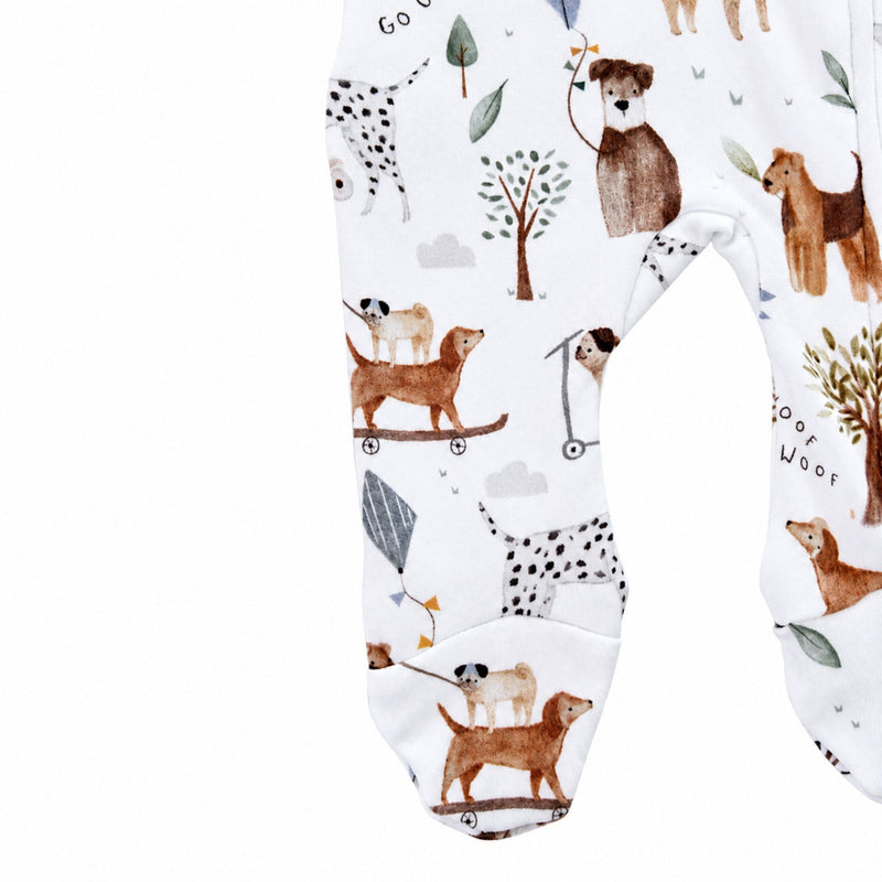 Close-up of the built-in footies on the Dogs Playground Footy Romper, showing brown dogs on skateboards, a spotted dog, trees, leaves and kite details on white cotton fabric.