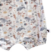 Close-up of a baby romper or bodysuit fabric with an all-over underwater print featuring whales, seahorses, coral, and sea urchins. A small brand tag is visible on the right side.