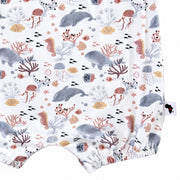 Close-up of a baby romper or bodysuit fabric with an all-over underwater print featuring whales, seahorses, coral, and sea urchins. A small brand tag is visible on the right side.
