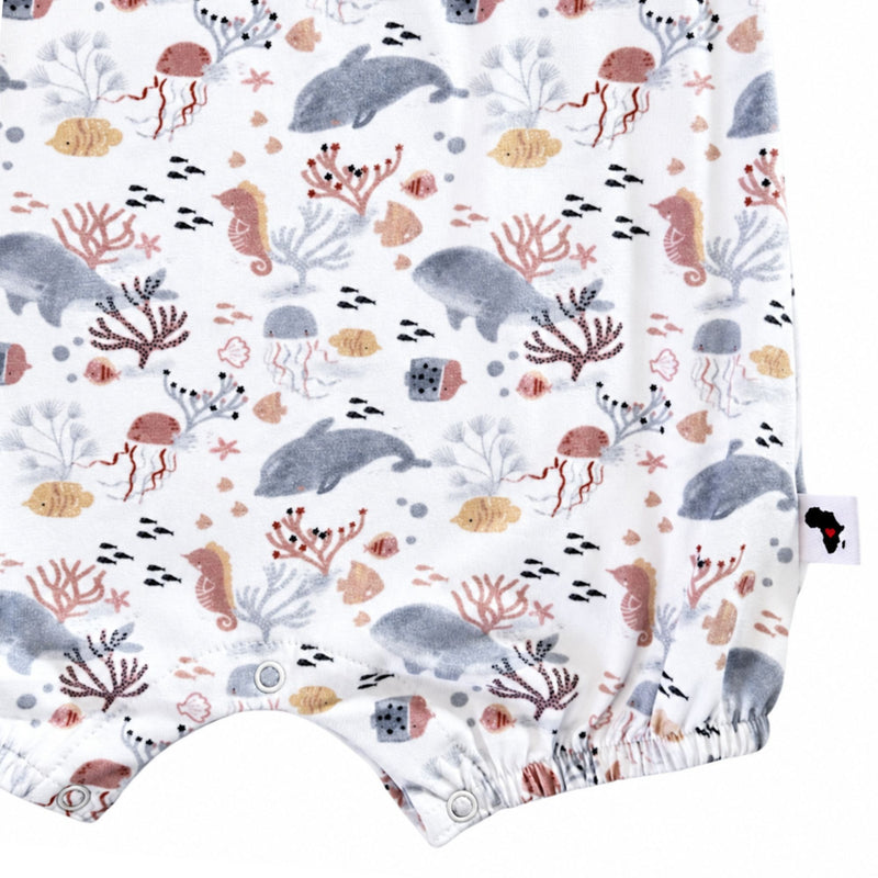 Close-up of a baby romper or bodysuit fabric with an all-over underwater print featuring whales, seahorses, coral, and sea urchins. A small brand tag is visible on the right side.