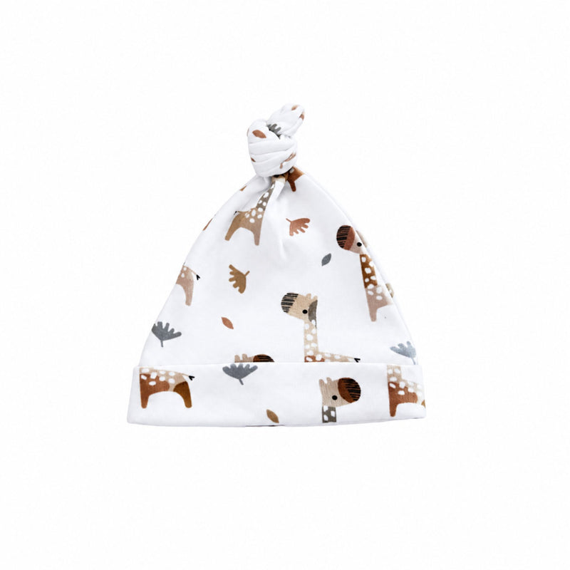 White baby knot beanie featuring playful brown, beige and grey giraffes with scattered leaves, a tied top knot and wide fold-over band.
