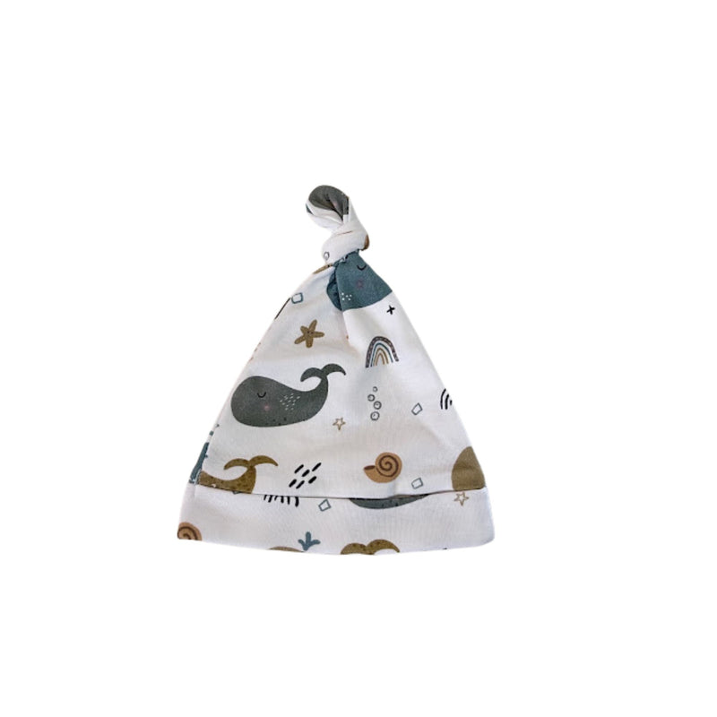 A baby beanie with a knotted top, featuring a white background with a whimsical pattern of a dark gray whale, a brown snail, a small rainbow, and scattered gold stars.