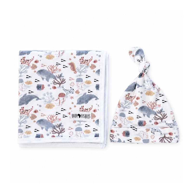 Folded white Finn and Friends blanket beside a matching knot beanie, with dolphins, seahorses, jellyfish and coral, white blanket edging and a turned-up beanie brim.