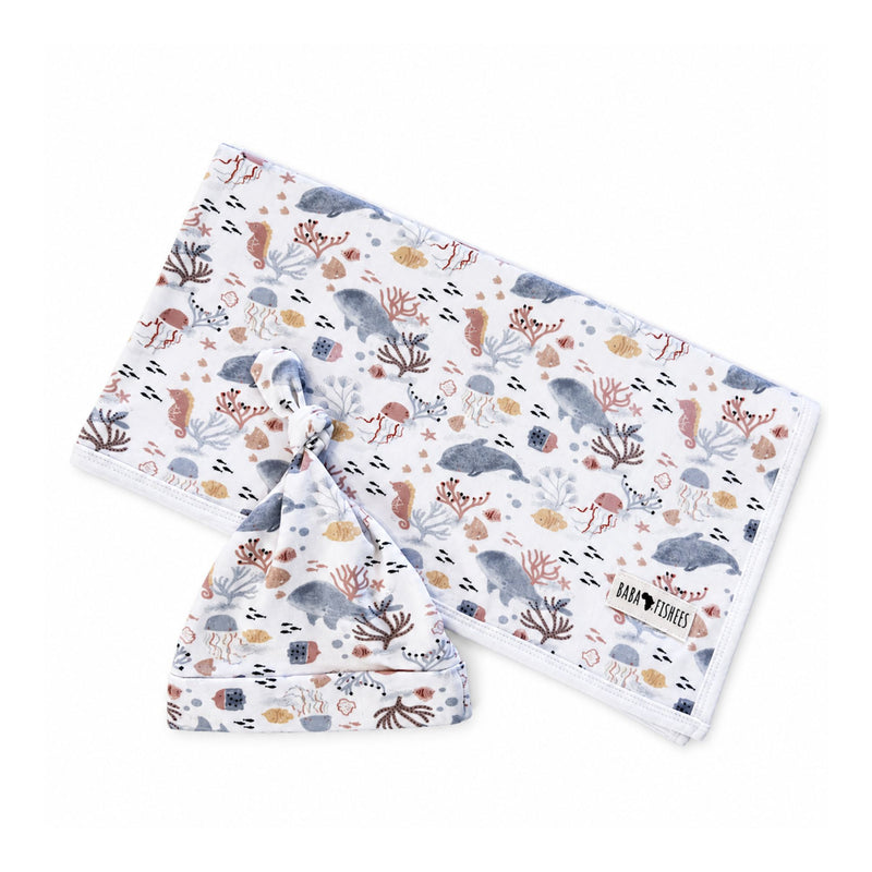 Finn and Friends blanket arranged diagonally behind a matching knot beanie, featuring blue-grey dolphins, pink sea creatures and golden fish on white fabric.