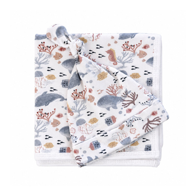 Finn and Friends knot beanie resting on a folded white baby blanket, both printed with blue-grey dolphins, pink seahorses, jellyfish and golden fish.