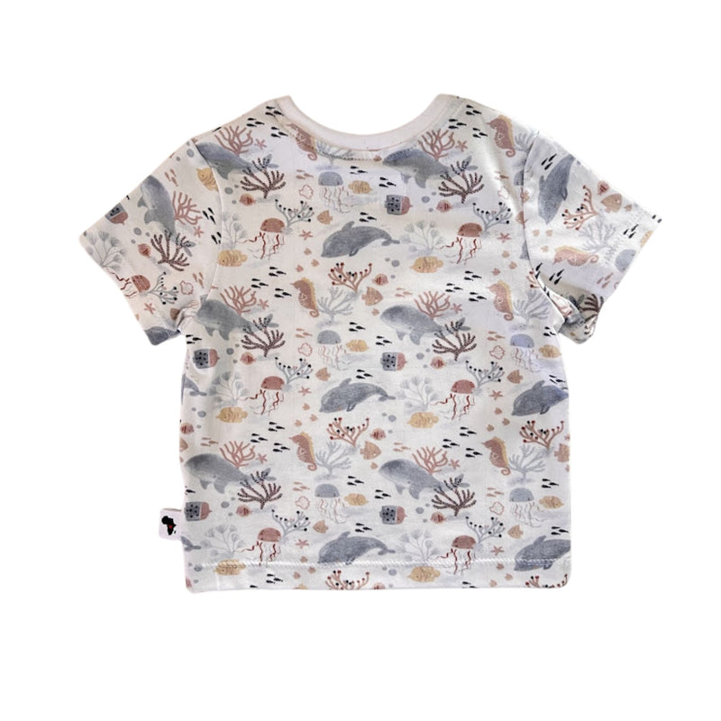 A short-sleeve children's t-shirt with an all-over whimsical print of sea creatures, including dolphins, seahorses, starfish, and coral, on a white background. 