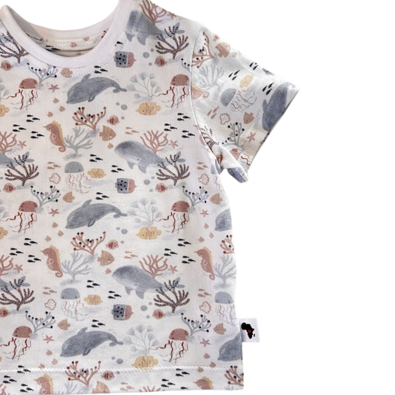 A short-sleeve child's t-shirt with an all-over underwater-themed print featuring whales, dolphins, jellyfish, seahorses, and coral in muted gray, terracotta, and beige tones.