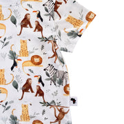 A close-up of a short-sleeve white children's T-shirt featuring a colorful safari animal print with illustrated monkeys, lions, zebras, toucans, and giraffes. A small fabric tag with an African continent silhouette is visible near the hem.