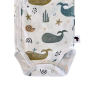 Bottom closeup of a white long-sleeve wrap-style baby onesie featuring a whimsical whale print in shades of blue, teal, and mustard, accented with rainbows, starfish, and seashells. Designed with press-stud closures for easy dressing.
