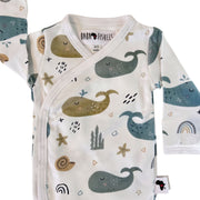 Neckline closeup of a white long-sleeve wrap-style baby onesie featuring a whimsical whale print in shades of blue, teal, and mustard, accented with rainbows, starfish, and seashells. Designed with press-stud closures for easy dressing.