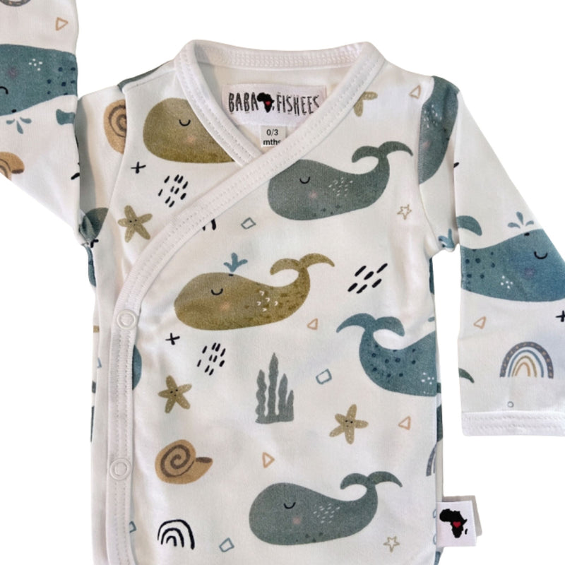 Neckline closeup of a white long-sleeve wrap-style baby onesie featuring a whimsical whale print in shades of blue, teal, and mustard, accented with rainbows, starfish, and seashells. Designed with press-stud closures for easy dressing.
