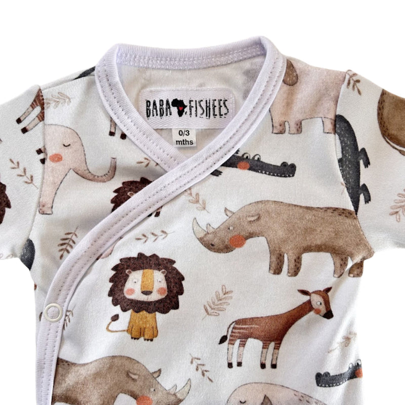 A close-up of a baby wrap-style romper with a white base and a playful safari animal print including a lion, rhinoceros, elephant, giraffe, and crocodile. The brand label 'BABA + FISHEES' is visible inside the collar.
