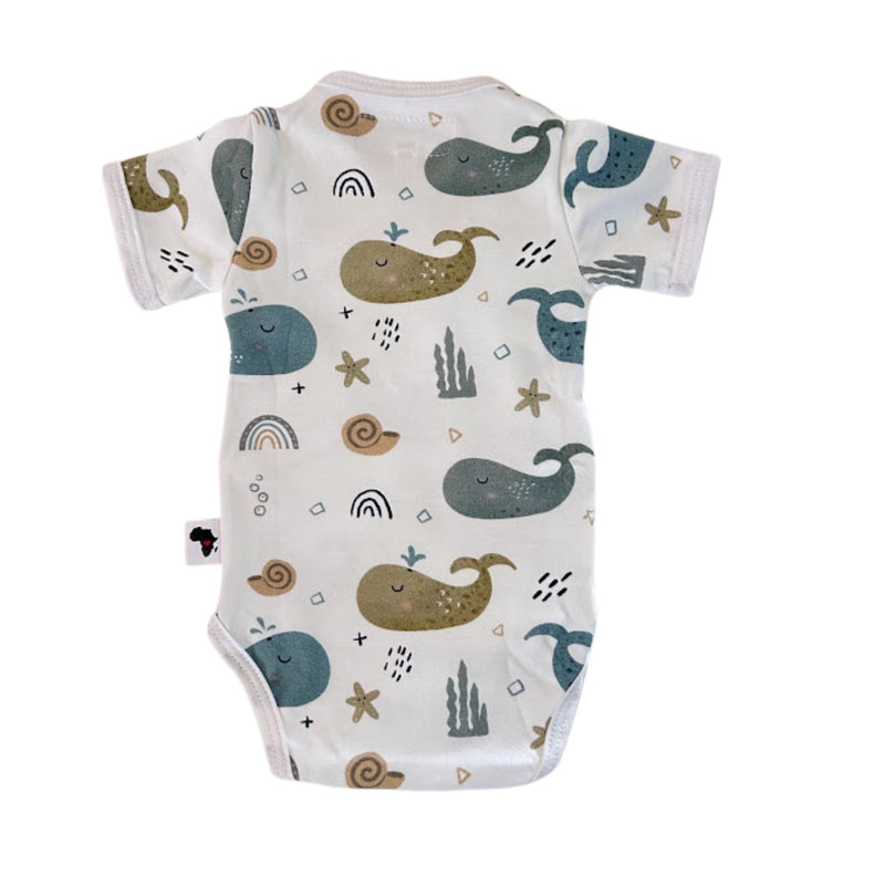 Back view of a white short-sleeve wrap-style baby onesie featuring a whimsical whale print in shades of blue, teal, and mustard, accented with rainbows, starfish, and seashells. Designed with press-stud closures for easy dressing.