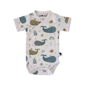 Front view of a white short-sleeve wrap-style baby onesie featuring a whimsical whale print in shades of blue, teal, and mustard, accented with rainbows, starfish, and seashells. Designed with press-stud closures for easy dressing.