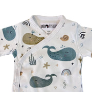Neckline closeup of a white short-sleeve wrap-style baby onesie featuring a whimsical whale print in shades of blue, teal, and mustard, accented with rainbows, starfish, and seashells. Designed with press-stud closures for easy dressing.